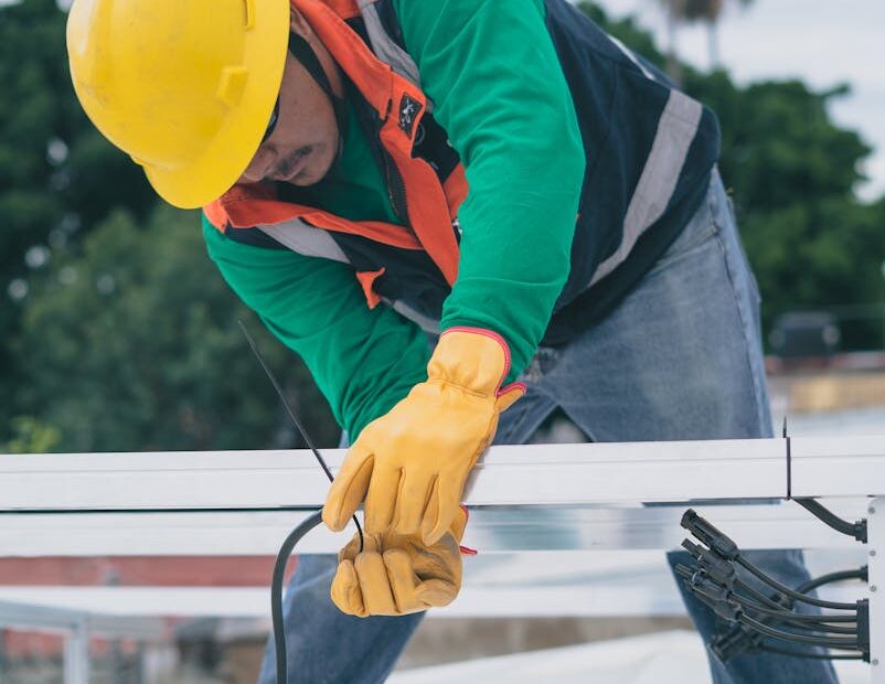 A construction worker wearing PPE installs electrical equipment on a roof.